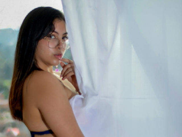 AnnieVolk with glasses, dark hair, looking sideways, touching her lip with her finger, white curtain, bare back, blue strap, slightly visible tits, soft natural light, calm expression.