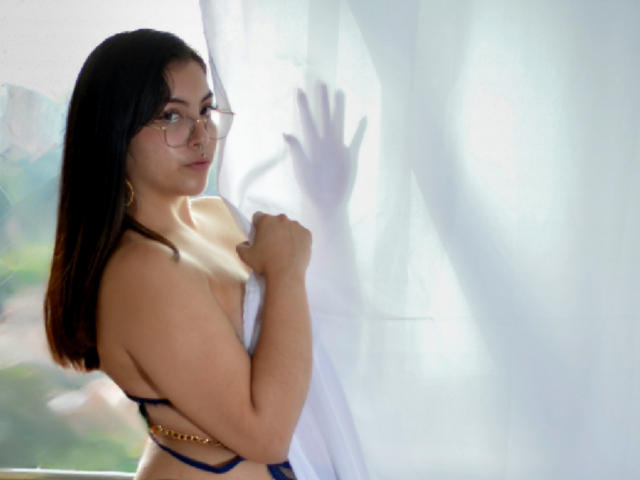 AnnieVolk with glasses, holding white curtain, showing back and side, wearing blue strappy top, hands on curtain, looking over shoulder, brown hair, light skin, subtle smile.