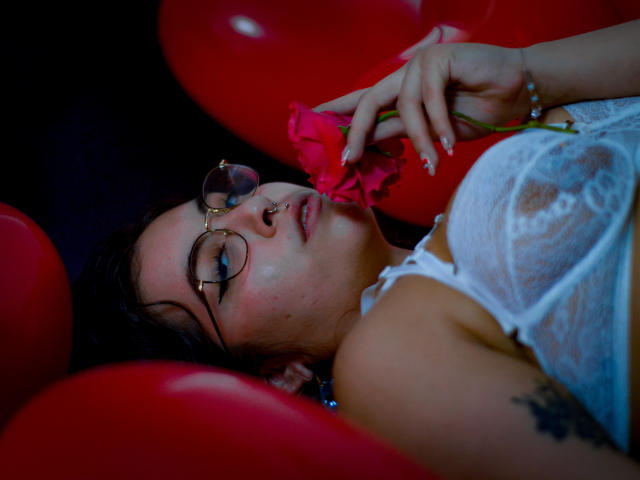 AnnieVolk con gli occhiali, un anello al naso, una camicetta di pizzo bianco, che tiene una rosa rosa, sdraiata su palloncini rossi, con un tatuaggio visibile sul braccio, che guarda verso l'alto, capelli scuri, trucco leggero, braccialetto..