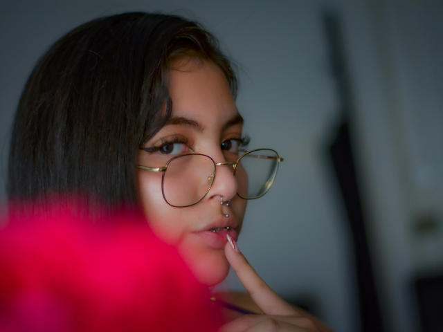 AnnieVolk with dark hair, gold glasses, nose ring, and lip ring, finger on bottom lip, blurry red flower in foreground. Her eyes are half-open, looking at camera.