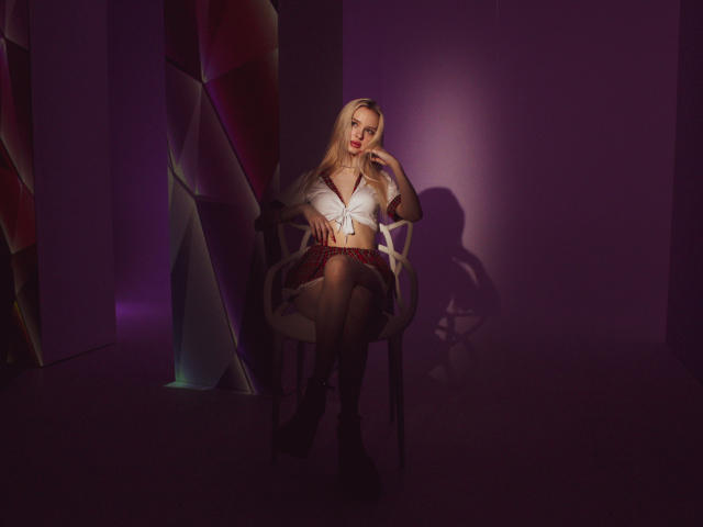 LexyGlam with long blonde hair, white tied-up top, black skirt, and boots, sitting on a chair, hand on chin, in dim purple light, shadow on wall.