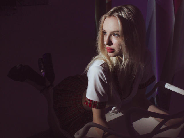 LexyGlam mit blonden Haaren, blauen Augen, rotem Lippenstift, weißem Hemd, rotem kariertem Rock, schwarzen Stiefeln, die an einem Stuhl lehnt, über die Schulter schaut, sexy, verführerisch..