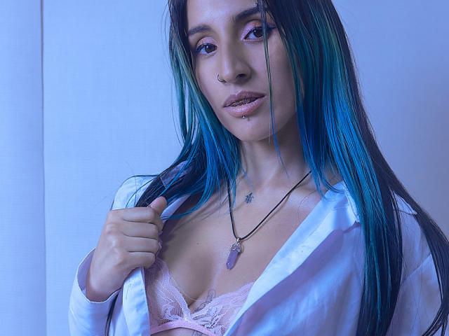 LeilaP con i capelli blu-verdi, un anello al naso e un reggiseno di pizzo rosa, indossa una camicia bianca aperta, tenendola con una mano, una collana con una pietra viola, e guarda la fotocamera..