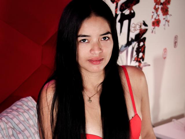 Paulette Leroy, met lang zwart haar, een rode bh en een subtiele glimlach, toont kleine borsten en een schattig gezicht, draagt een sterrenketting en zit op een bed..