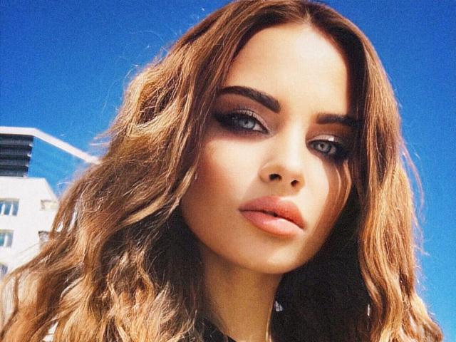 CharmingVixeny heeft golvend bruin haar, volle lippen met een roze lippenstift, en opvallende blauwe ogen met donkere eyeliner. Ze kijkt iets naar beneden, met een helderblauwe lucht op de achtergrond..
