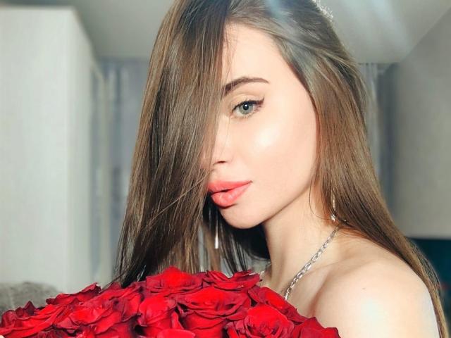 Uma CharmingVixeny com longos cabelos castanhos cobrindo um olho, segurando um buquê de rosas vermelhas, tem pele brilhante e lábios rosados, usando um colar de prata, e parece sedutora..