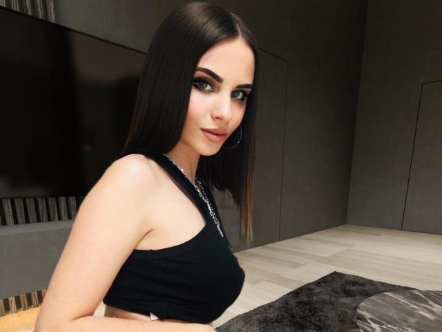 CharmingVixeny con cabello castaño largo, ojos verdes y aros grandes, vistiendo una camiseta negra sin mangas, mostrando sus hombros desnudos y pequeños senos, en una habitación con poca luz..