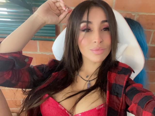 LeilaP con cabello largo y castaño, párpados rosados, camisa de cuadros rojos, sujetador de encaje rojo, mostrando sus pechos, mano en el cabello, labios rosados, collar, silla blanca, cabello azul en el fondo..