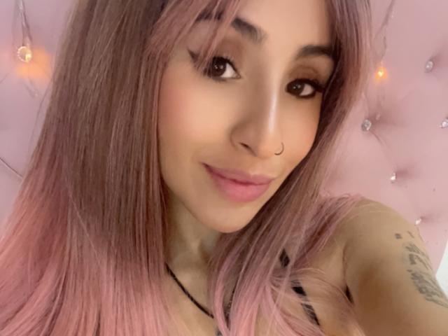LeilaP, con cabello rosa, aro en la nariz y delineador de ojos, sonriendo a la cámara, mostrando tatuajes en su brazo derecho, vistiendo una blusa negra, iluminación suave, fondo rosa..