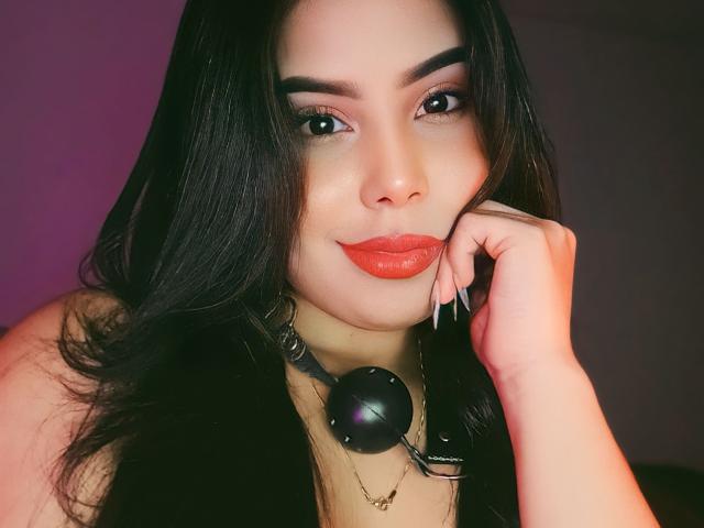 EvaxRose met donker haar, rode lippenstift en een zwarte ball gag halsband, lachend, haar wang aanraakend, haar glanzende roze nagels showend, en een zwart mouwloos topje dragend..