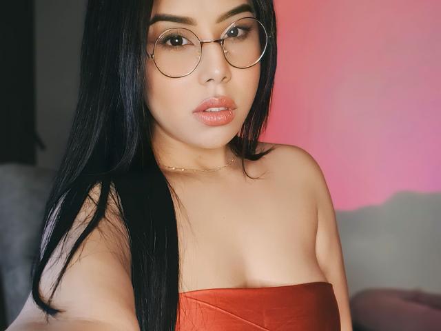 EvaxRose met lang zwart haar, die ronde brillen draagt, een rode top zonder bandjes, en een gouden ketting, waarbij haar borsten zichtbaar zijn, kijkt naar de camera met glanzende, roze lippen..