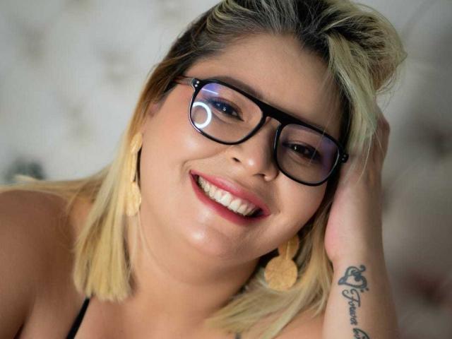 MilenaDutty, com cabelo loiro, óculos pretos, sorrindo, usando brincos com franjas, com a mão no cabelo, tatuagem no pulso, olhando para a câmera. Seios parcialmente visíveis, mostrando um visual fofo e sexy..