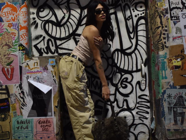KeilaPalaciosX, avec ses cheveux foncés, portant des lunettes de soleil, un débardeur rose et un pantalon cargo jaune, appuyée contre un mur recouvert de graffitis. Ses fesses sont visibles, et elle a un tatouage..