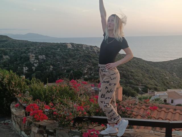 Saveta staat met haar arm omhoog, ze draagt een zwarte top en een broek met een patroon, witte sneakers, op een dak met rode bloemen, haar blonde haar wappert in de wind, met uitzicht op de kust..