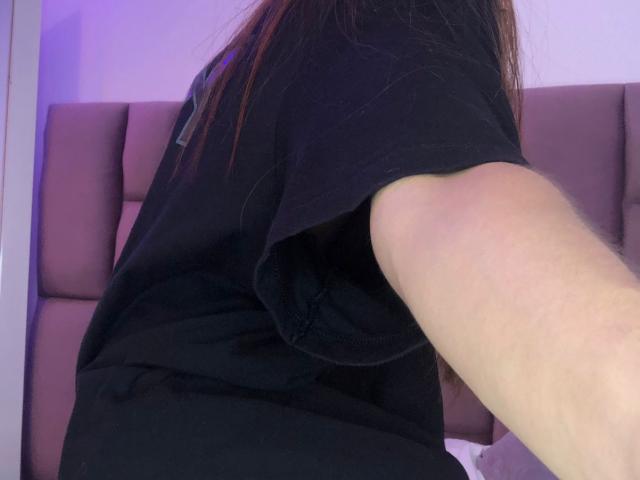Das schwarze Hemd von CieloSmith, das einen Teil ihres Arms und Oberkörpers zeigt, mit langen braunen Haaren, in einem violett beleuchteten Raum..