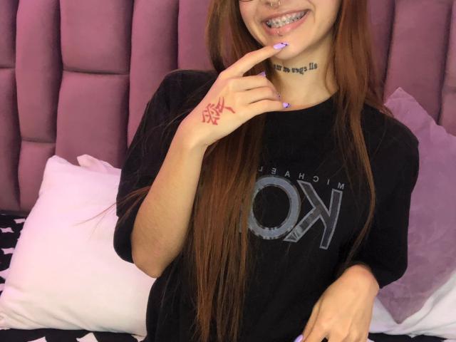 CieloSmith mit langen braunen Haaren, schwarzem T-Shirt, rosa Nagellack, Finger-Tattoo, Nasenring (Septum), lächelnd, mit sichtbaren Zähnen, weißes Kopfkissen, violette Kopfteil und schwarze Bettdecke im Hintergrund..