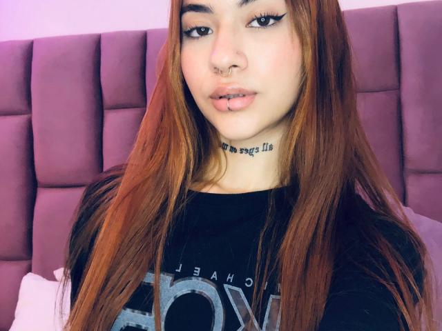 CieloSmith, con cabello rojo largo, delineador negro, piercing en la nariz, piercing en el labio, collar, camisa negra, mirando a la cámara, cabecera morada en el fondo..