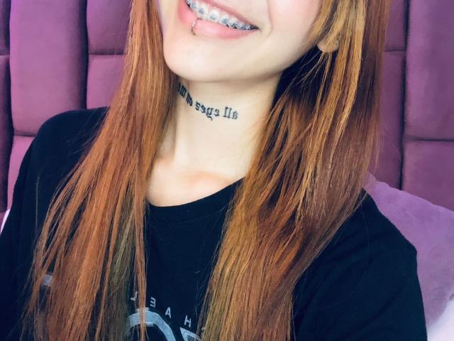 CieloSmith with long red hair, black shirt, metal teeth, and a tattoo on her neck, smiling. Purple, cushioned background.