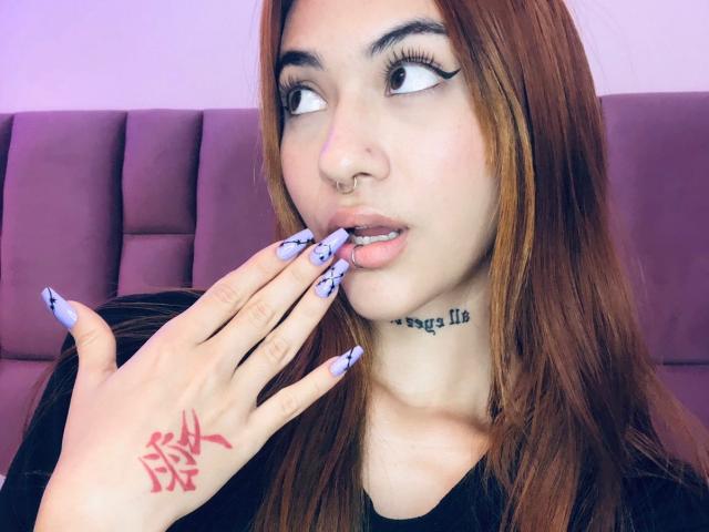 CieloSmith mit hellbraunen Haaren, schwarzem Eyeliner, Nasenring, tätowierter Hand mit blauen und weißen Nägeln, die Lippen berührend, einem violetten, gepolsterten Hintergrund und einem schwarzen Oberteil..