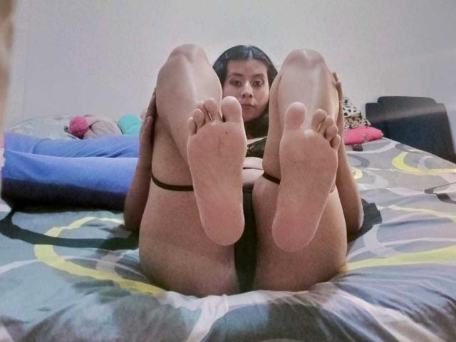 PaquitaHot está acostada en una cama con las piernas levantadas, mostrando las plantas de sus pies y su vulva. Lleva ropa interior negra, tiene el pelo oscuro y una expresión neutra. Sus pies están en primer plano..