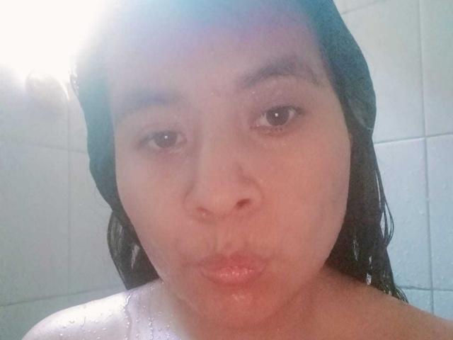 PaquitaHot tiene el cabello negro y húmedo, ojos marrones y labios carnosos, con gotas de agua en sus hombros desnudos en un baño, mirando directamente a la cámara..