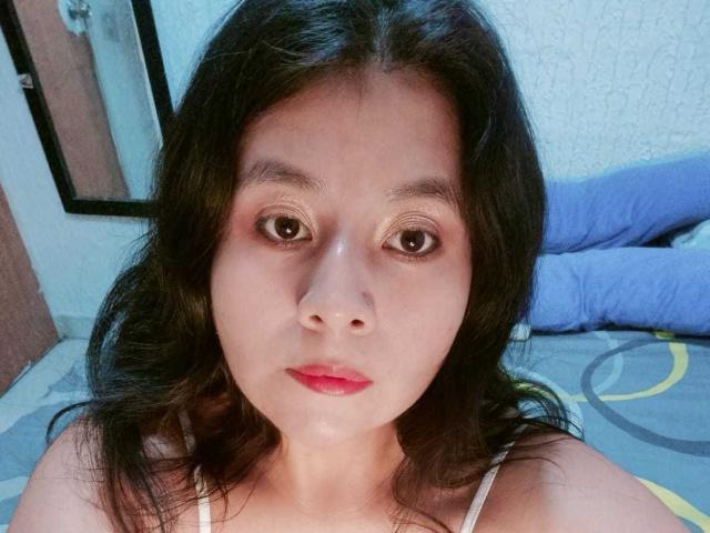 PaquitaHot a de beaux yeux, des cheveux noirs ondulés et du rouge à lèvres. Elle porte un débardeur blanc et regarde directement l'appareil photo avec une expression neutre..