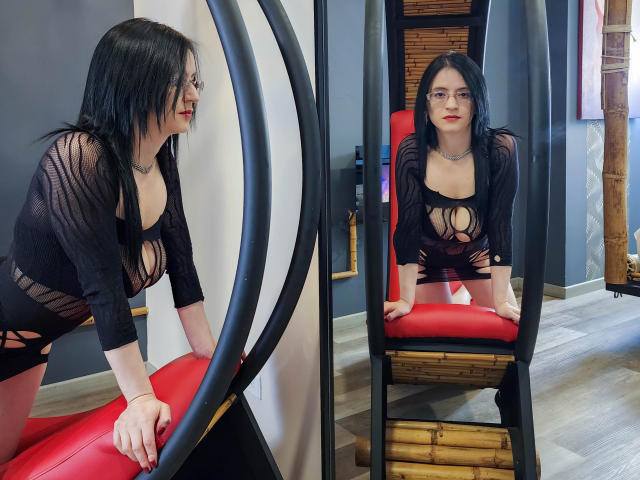 AlanaMoneth with black hair, glasses, and red lipstick, wearing a revealing black mesh top with cut-out breasts, leaning on a red and black exercise machine. Mirror reflection.