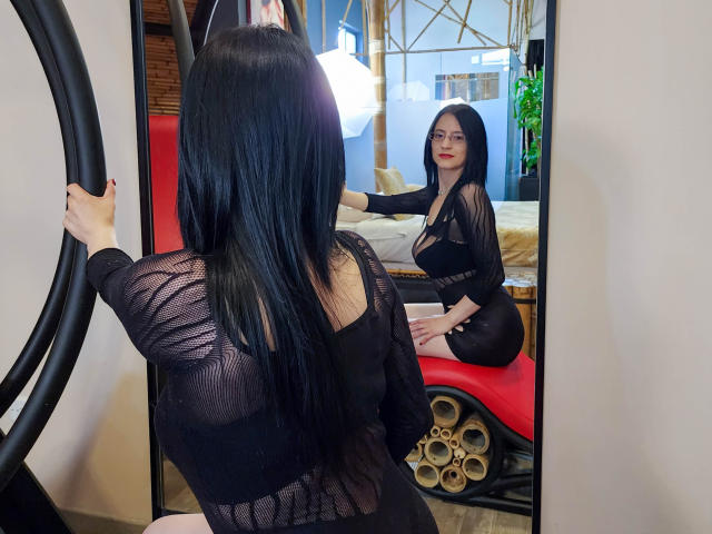 AlanaMoneth with long black hair, sheer black top, and black dress, sitting on red bench, reflecting in mirror, showing her ass and small tits. Mirror captures her front.