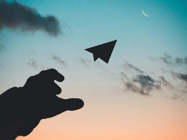 Silhouette de la main de SuperbissimaTroiana, avec les doigts légèrement recourbés, lâchant un avion en papier dans un ciel de coucher de soleil avec une lune croissante..