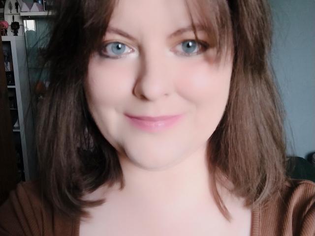 DahliaGray tem olhos azuis, cabelo castanho claro na altura dos ombros e um sorriso discreto com lábios rosados. Ela está vestindo uma blusa marrom, e seu nariz é furado..