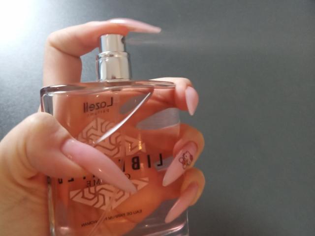 Zanoza's hand, met lange, roze nagels, houdt een fles roze Hsosl nagellak vast. Haar vingers houden de fles vast bij de pomp. Haar pinknagel heeft een klein hartje-design..