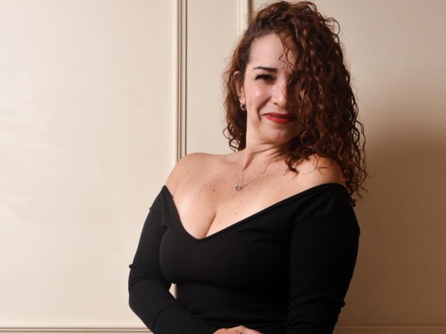 ScarlettthVega with curly brown hair, black off-shoulder top, showing her tits, smiling, red lipstick, necklace, arms crossed, standing against beige wall.