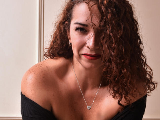 ScarlettthVega, con cabello rojo rizado, sonriendo, vestida con una blusa negra con un hombro descubierto, mostrando sus pechos, hombros con pecas y un collar de plata con forma de corazón. Su lápiz labial rojo complementa su apariencia segura..