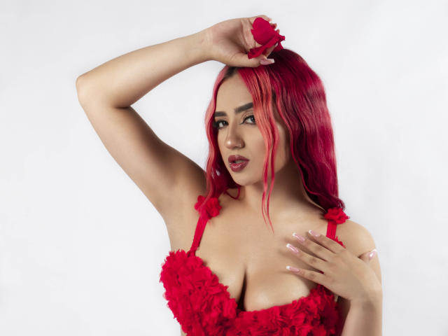 Melanie Kane, com cabelo rosa, vestido floral vermelho e unhas rosa, segurando uma flor vermelha acima da cabeça, mostrando os seios, olhando para a câmera com um leve sorriso..