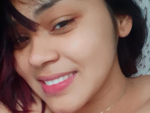 El rostro de IntiAmor, sonriendo con lápiz labial rosa, ojos marrones, cabello oscuro con puntas moradas y una nariz pequeña, lleva un collar de oro delgado, en una foto de primer plano..