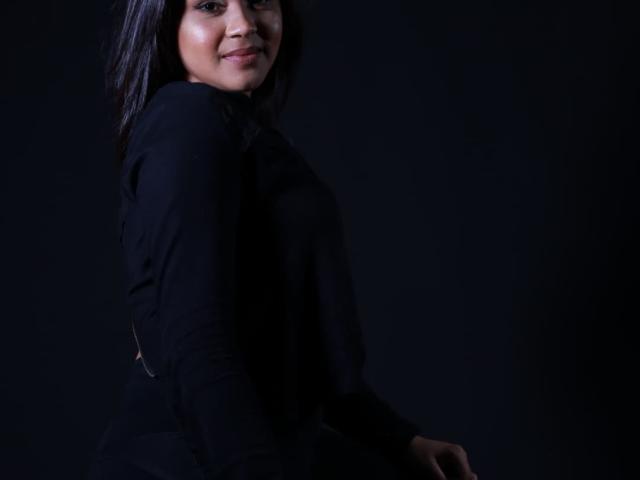 IntiAmor, una mujer de cabello oscuro, sonriendo, vestida con una blusa negra de manga larga, mirando a la cámara por encima del hombro, fondo negro, luz sutil en el lado izquierdo, manos sobre el asiento, segura de sí misma, atractiva..