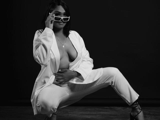 IntiAmor with dark hair, wearing a white open shirt, exposing large tits, white pants, and laced shoes, crouching, holding glasses, smiling, black background.