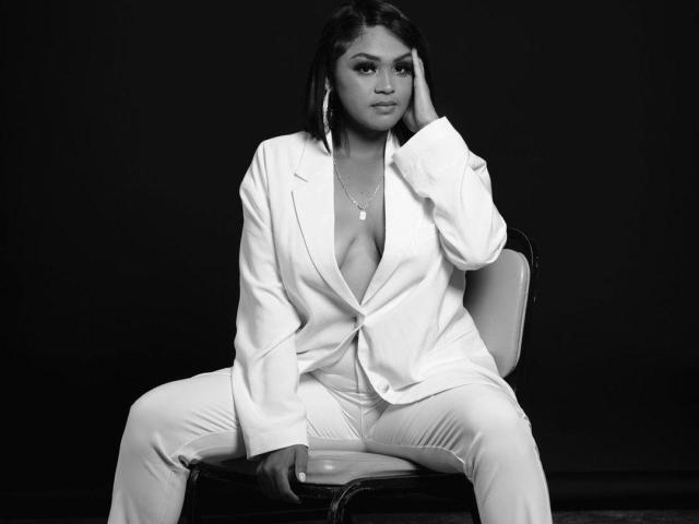 IntiAmor sits on a chair, legs spread, wearing an open white shirt revealing her tits, hand on her head, looking at the camera, black background, black and white.
