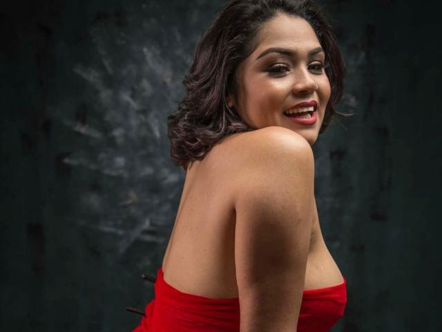 IntiAmor, com cabelos escuros e ondulados e batom vermelho, sorrindo para a câmera, vestindo um vestido vermelho sem alças, mostrando seus ombros e parte superior das costas. Seus seios estão parcialmente visíveis..