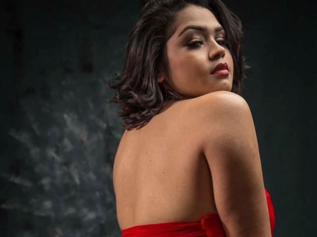 IntiAmor with dark wavy hair, red strapless dress, looking back, showing bare shoulders and upper back, with a sultry expression, dark green background.