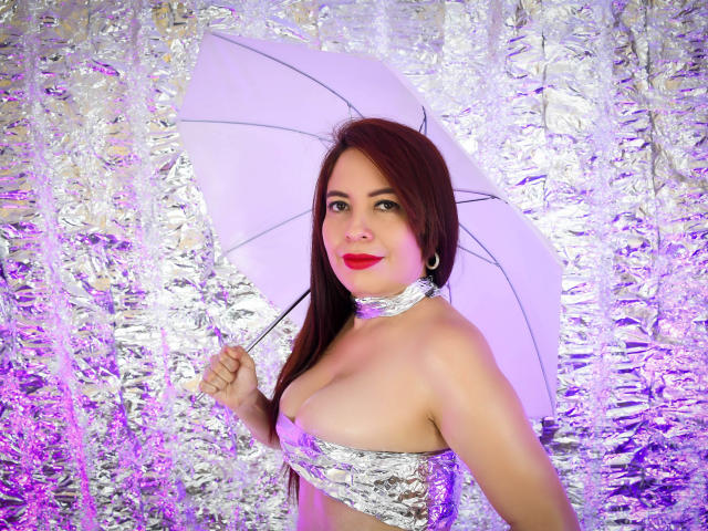 Esperanza Castillo, con cabello rojo, sosteniendo un paraguas blanco, vestida con papel de aluminio plateado cubriendo sus pechos y cuello, luz púrpura, sonriendo, con lápiz labial rojo, en una fotografía..