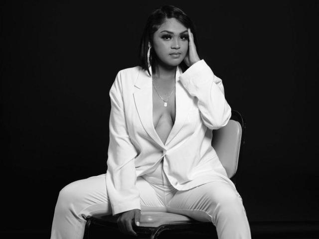 IntiAmor, con piel oscura, vestida con un blazer y pantalones blancos, está sentada en una silla, con la mano en la mejilla, mostrando sus grandes senos, fondo negro, con una expresión seria, lleva un collar, foto en blanco y negro..