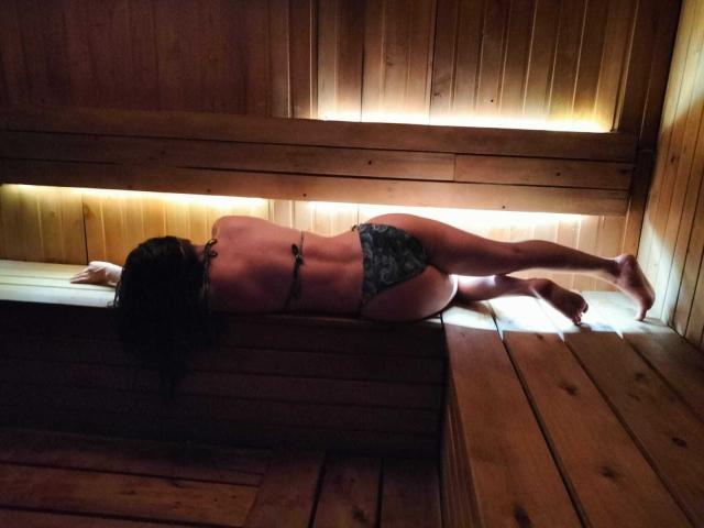 Contelaya, lying face-down in a wooden sauna, wearing a black bra and floral panties, exposing her bare back and ass. Her legs are stretched out, and her head is down.