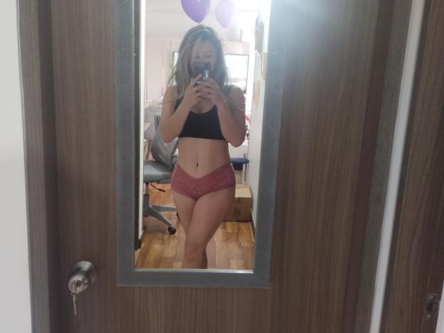 Contelaya, met blond haar, draagt een zwarte sportbeha en roze kantonderbroek, en maakt een spiegelzelfportret, waarbij haar borsten en billen zichtbaar zijn, in een kamer met houten vloeren..