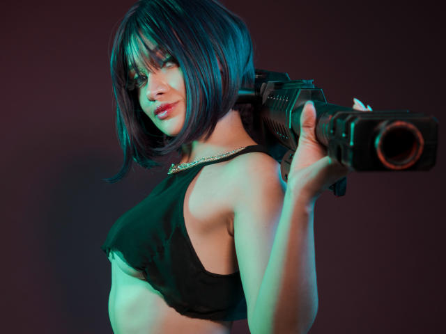 AngelCutie avec une coupe de cheveux noire courte, tenant une arme à droite, portant un crop top noir, montrant ses seins, avec un regard confiant et sexy, et un éclairage vert-bleu..