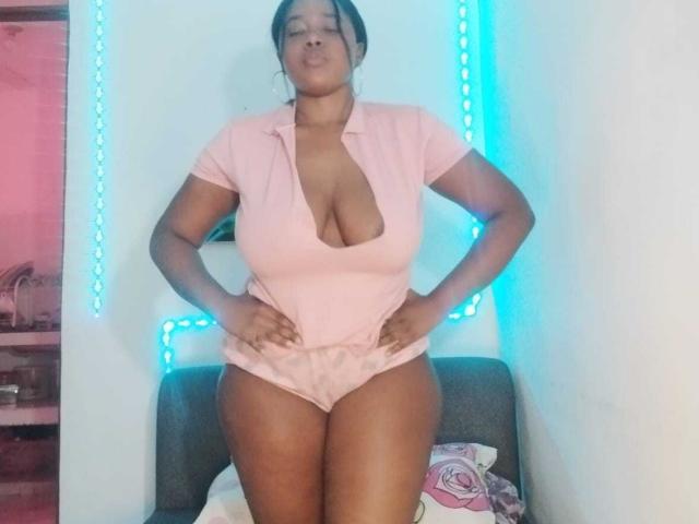 EmiraHot with dark skin, wearing a pink, low-cut, short-sleeve top, showing large, round tits, and pink panties, standing with hands on hips, blue LED strip behind.