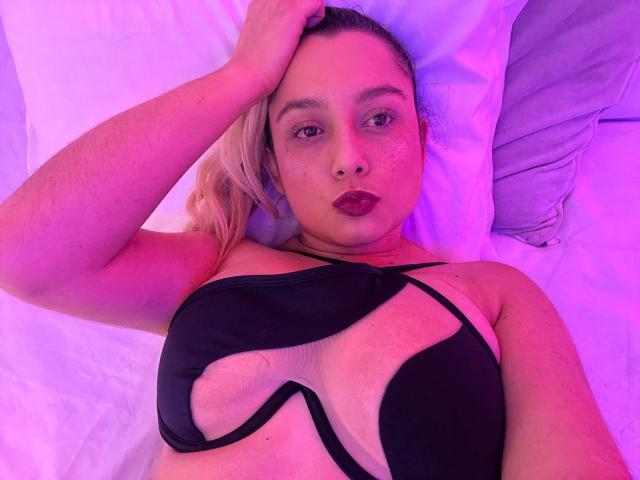 AmayaJhons est allongée sur un lit, vêtue d'un soutien-gorge noir avec découpes, qui expose ses seins. L'éclairage est rose-violet. Elle a une main dans ses cheveux blonds, ses lèvres rouges sont pincées, et elle regarde l'appareil photo..