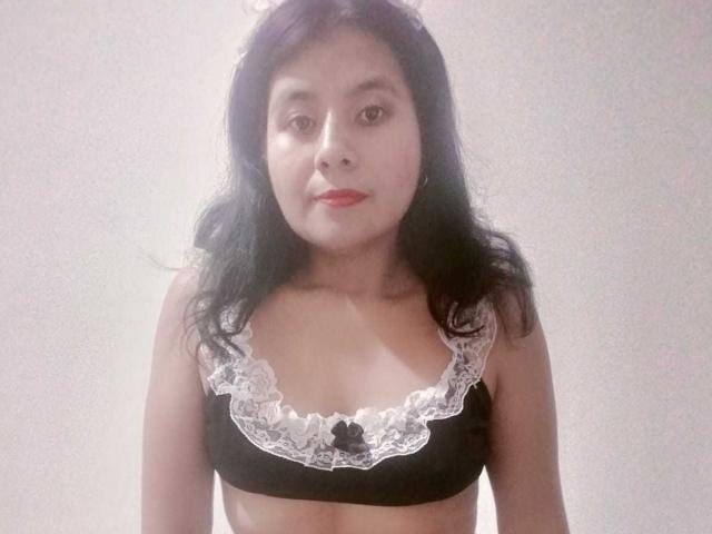 Una mujer madura, atractiva y sensual, con cabello negro, gafas y medias negras hasta el muslo, acostada en una cama con sábanas rojas y con motivos florales, mostrando sus pechos y vulva, con un dedo en la boca, y un teclado cerca..
