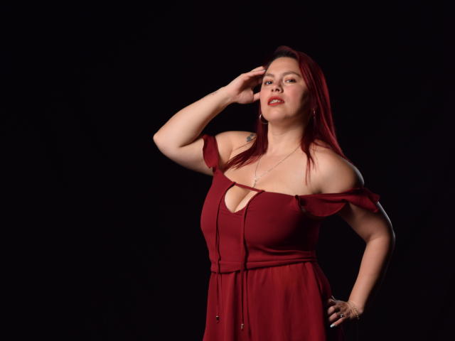 SalmaBrown con un vestito rosso, a spalle scoperte, una mano sulla testa, seno di media grandezza, pelle chiara, capelli rossi, sfondo nero, posa sicura, leggero sorriso, tatuaggio discreto sulla spalla sinistra..