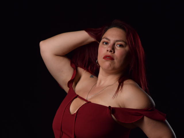 SalmaBrown with red hair, wearing a red off-shoulder dress, has a hand in her hair, showing a small tattoo on her right shoulder and a silver necklace.
